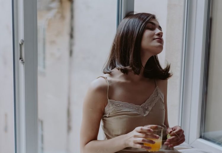 Woman in a spaghetti strap dress savoring orange juice on a window sill, indoors.