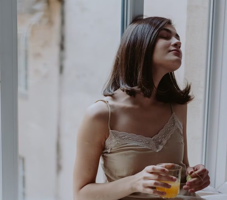 Woman in a spaghetti strap dress savoring orange juice on a window sill, indoors.